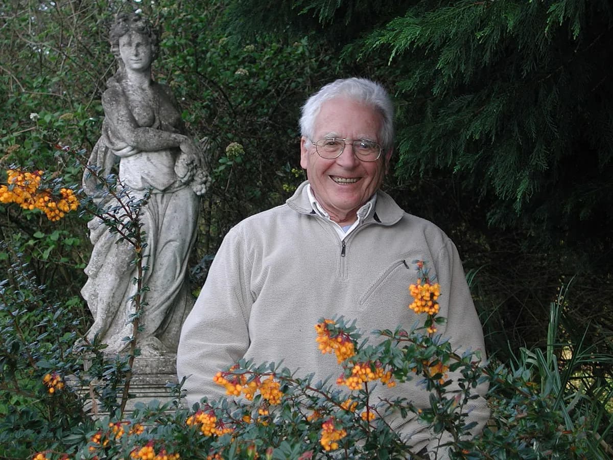 James Lovelock smiling in a garden, beside a stone statue and a shrub of orange berries.