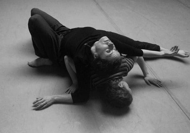 Two dancers in black balanced on the floor together in a contact improvisation duet.