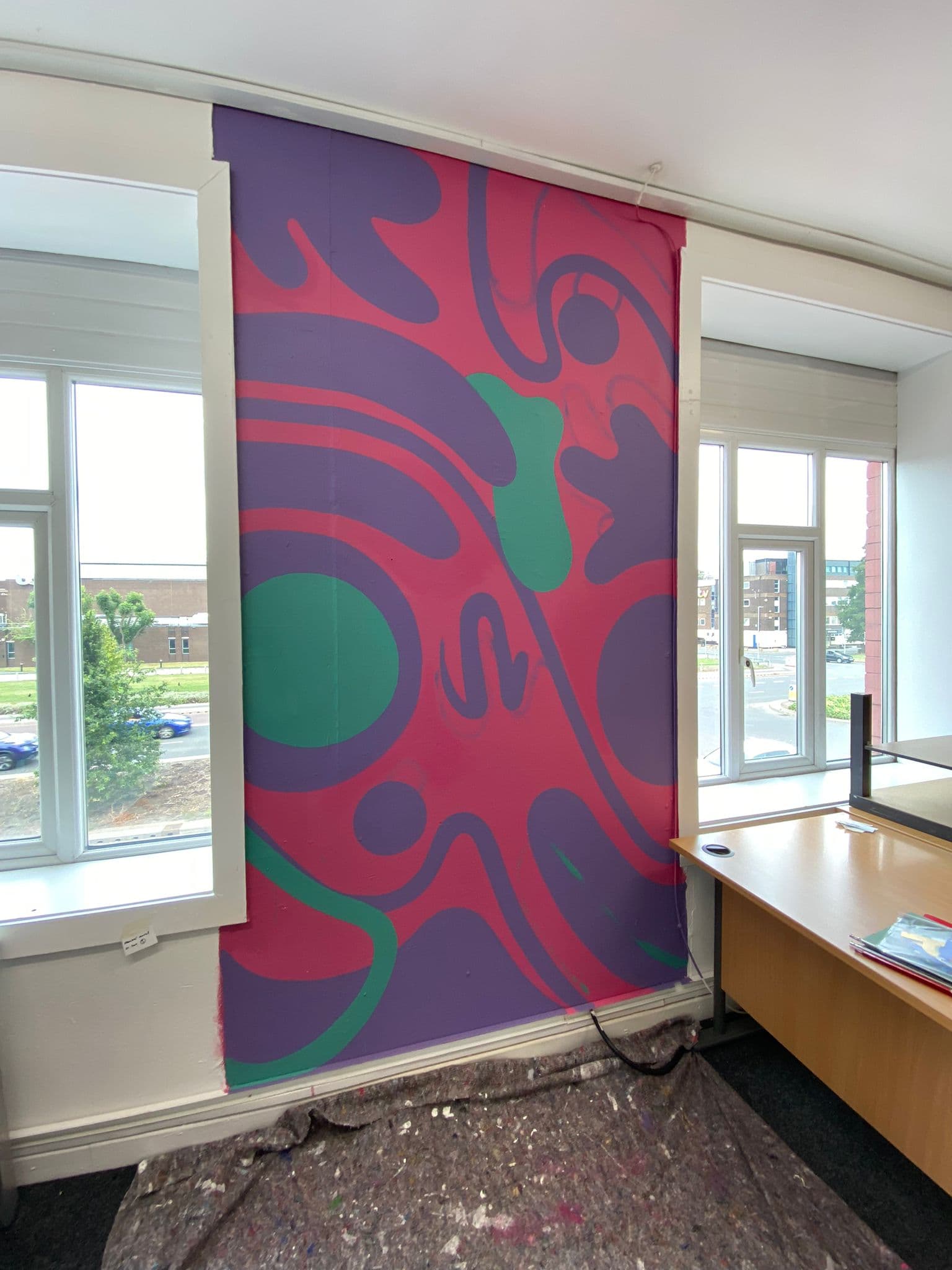A large pink, purple and green abstract mural painted on a studio wall between two windows.
