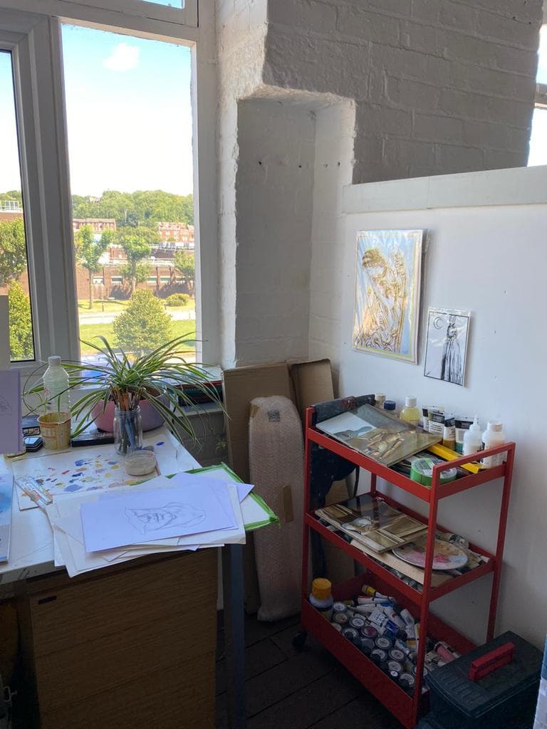 A studio corner with a paint-covered desk by the window and a red trolley loaded with paint tubes.
