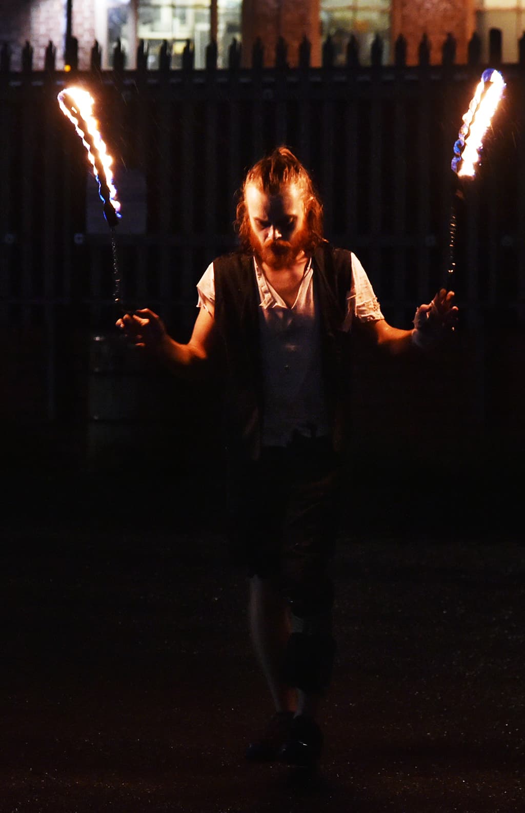 A fire performer holding chains attached to two burning props at night.