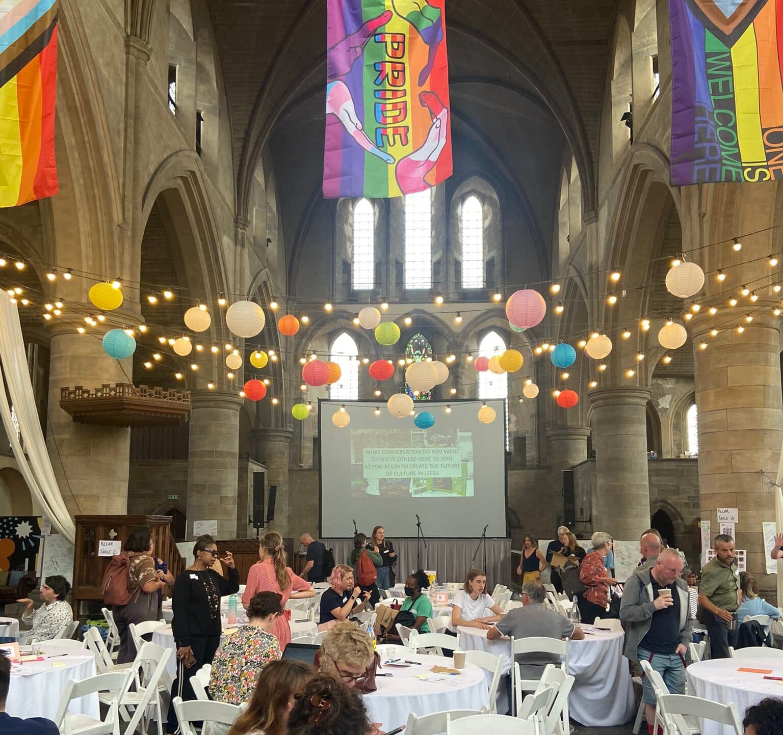 People talking around tables beneath paper lanterns and Pride flags at Left Bank Leeds.