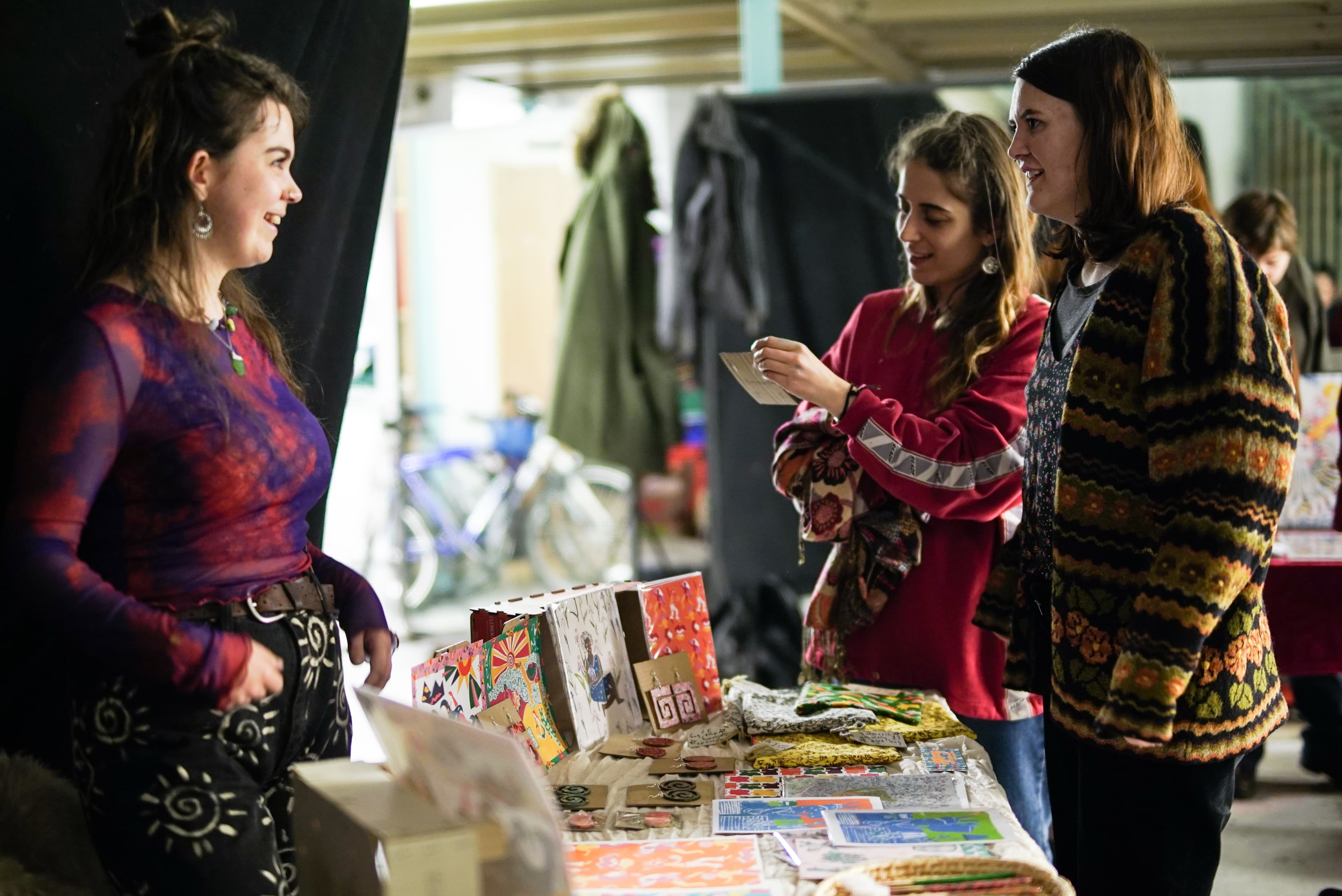 A maker talking with two visitors across her stall of handmade cards, earrings and textiles.