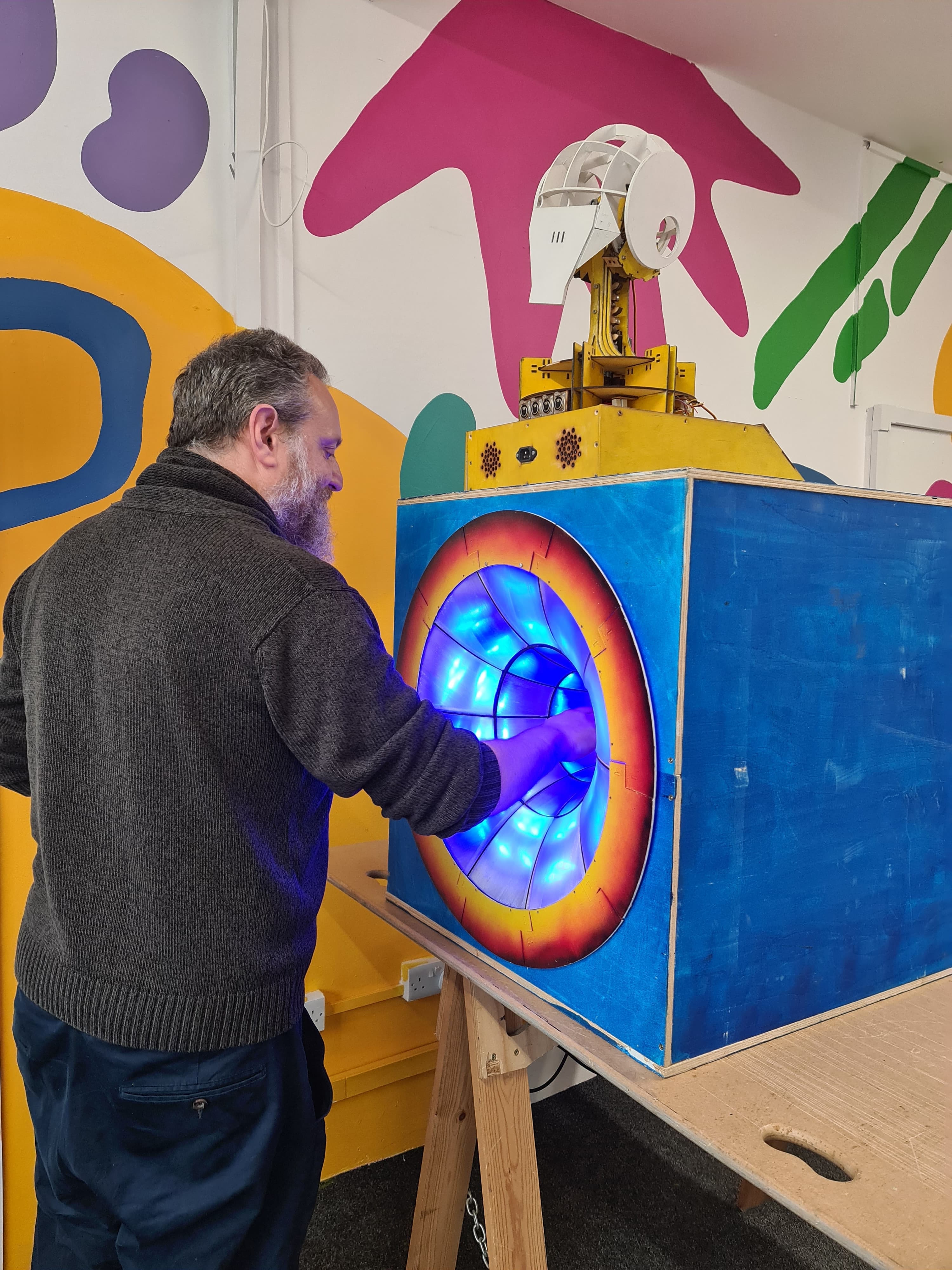 A person interacts with a brightly lit, blue-and-orange art installation at Open Source Arts. Their hand is inside a glowing tunnel of blue LED lights built into a painted wooden box, topped with a quirky yellow-and-white mechanical structure.