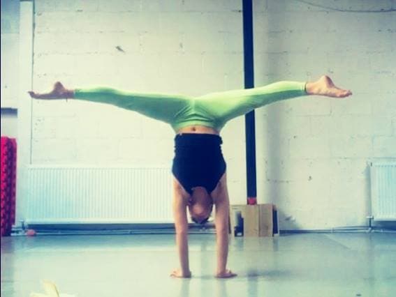 A performer holding a straddle handstand in front of a white brick wall.