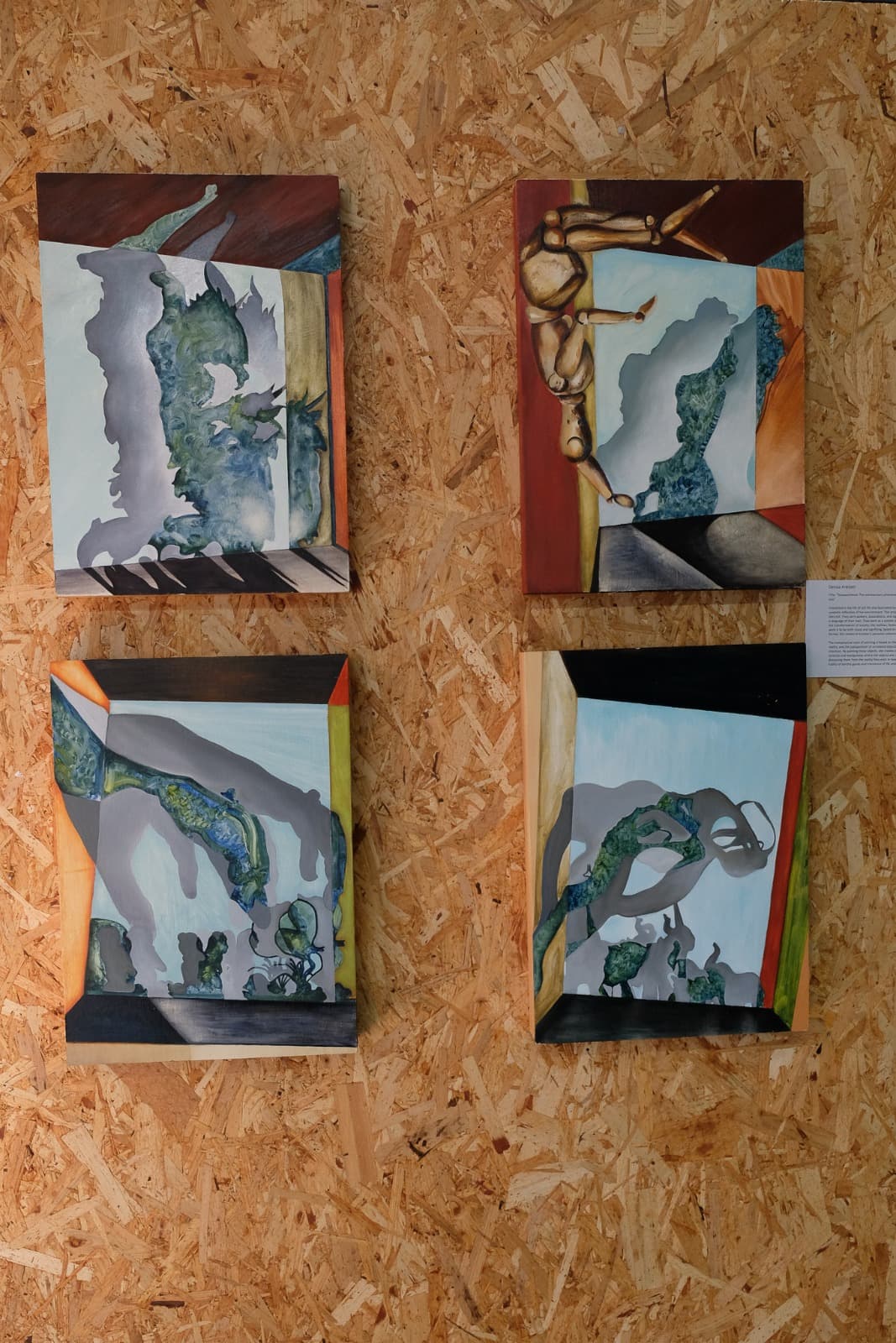 Four surreal paintings displayed together on a wooden chipboard wall at Open Source Arts. Each canvas features abstract organic shapes in muted tones of blue, grey, and green, set within geometric frames of warm reds, yellows, and oranges. One painting includes a wooden mannequin figure in an unusual pose, blending the surreal with the figurative.