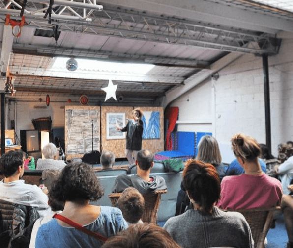An audience seated on sofas and chairs watching a performer on a small stage in the warehouse venue.