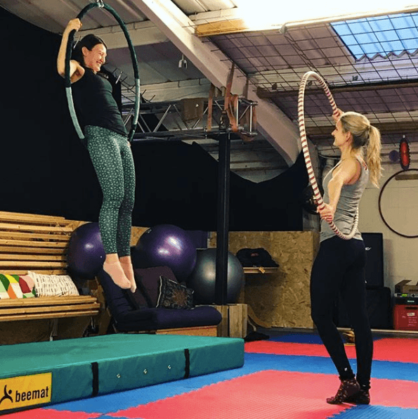 Two people training in the aerial space, one hanging from an aerial hoop, one holding a hula hoop.