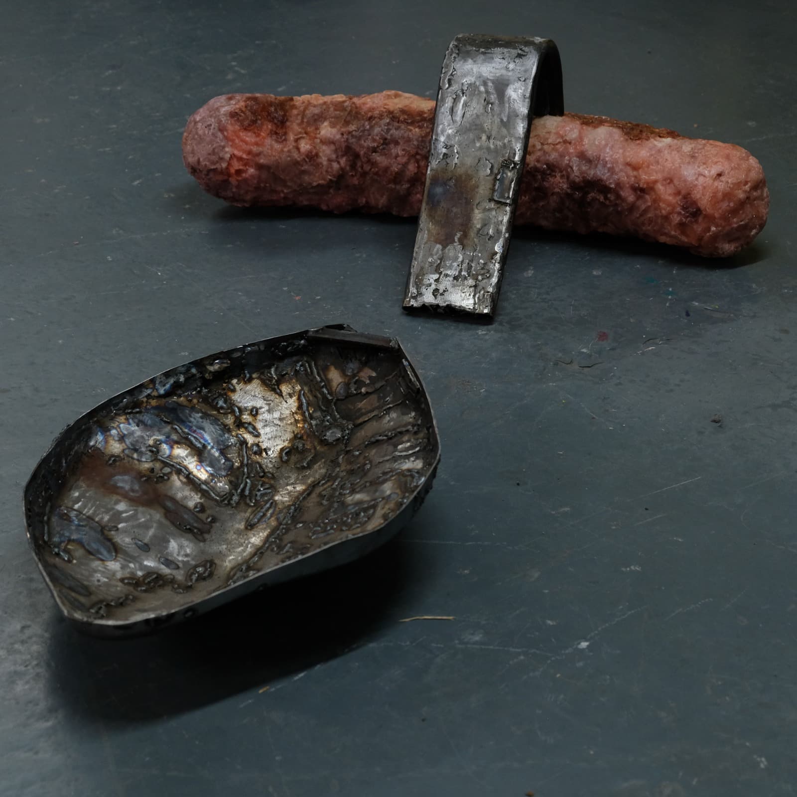 Two small sculptural objects placed on a dark grey floor. One is a pink, organic-looking cylindrical form with a rough surface, partially clasped by a curved strip of welded metal. The other is a shallow, irregularly shaped metal bowl with a scorched and textured surface, showing heat marks and welding scars.