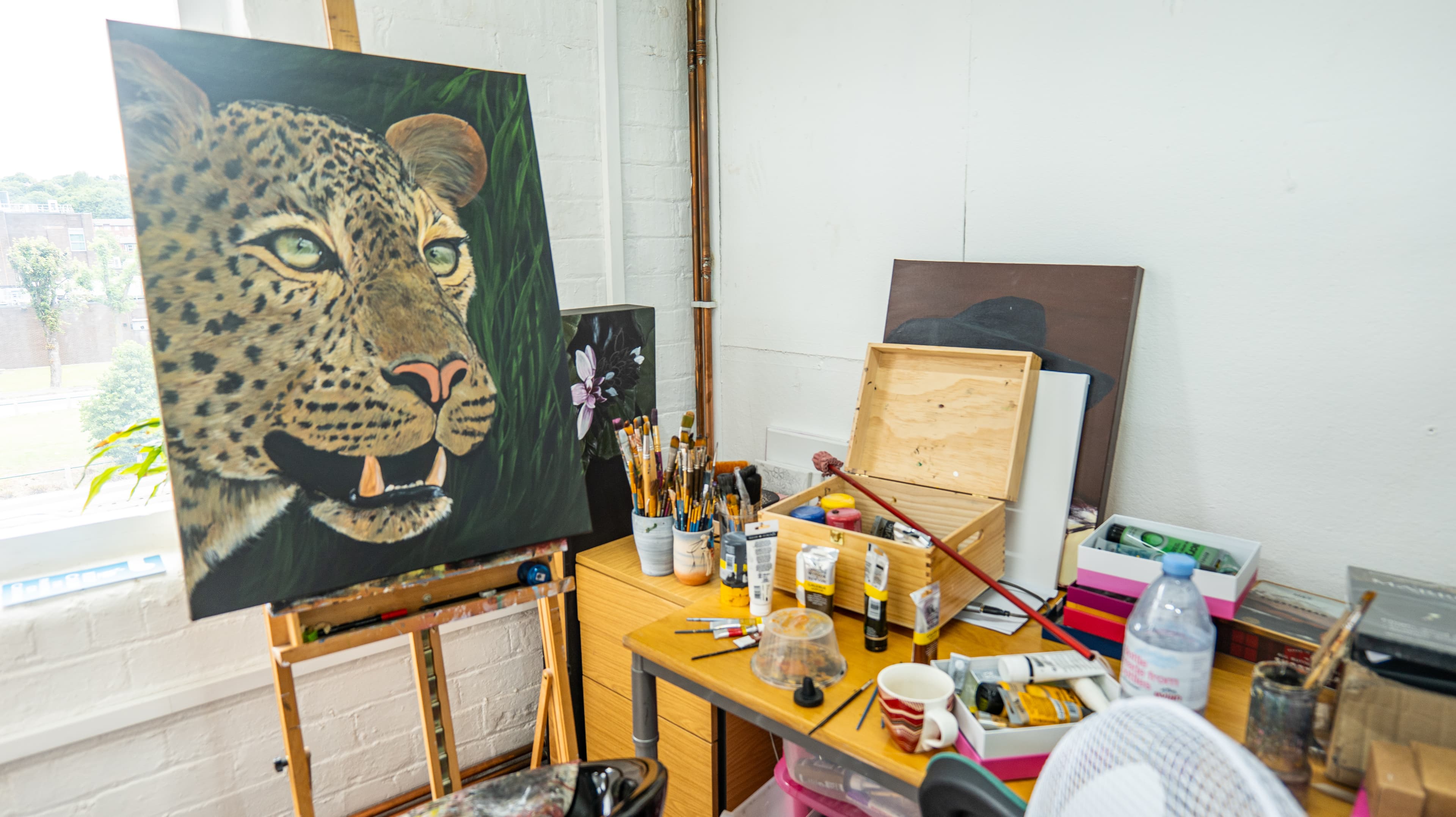An artist’s studio with a large painting of a leopard on an easel. The desk beside it is cluttered with art supplies, including paint tubes, brushes, jars, a wooden box, a striped mug, and a bottle of water. Other canvases lean against the wall, including one with a flower and another with a figure wearing a hat. Natural light enters through a nearby window.