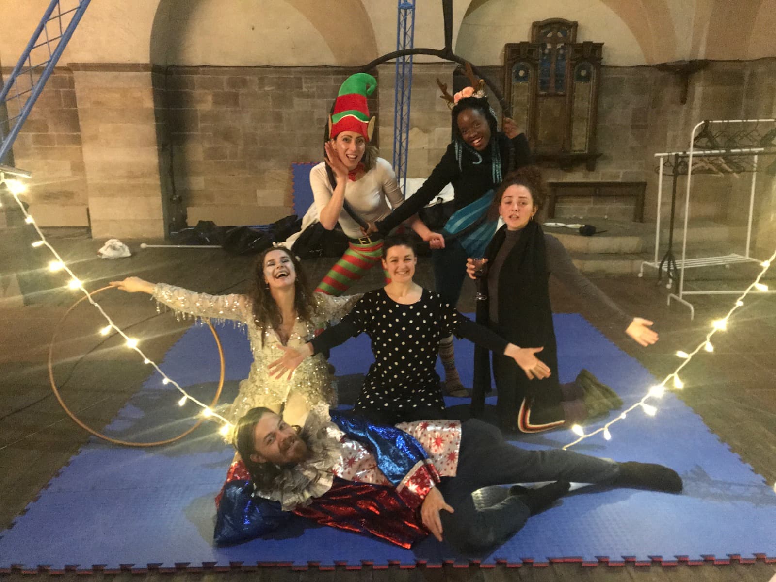 Six performers in costume posing on blue foam mats, with hoops and fairy lights.