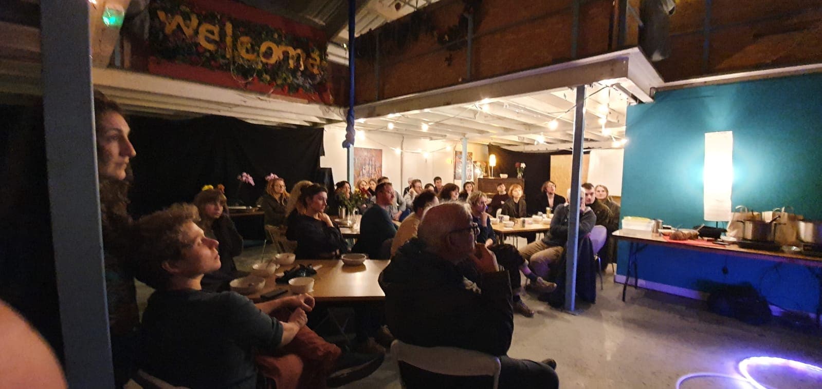 The Art SOUP audience with their soup bowls, watching a pitch under a handmade Welcome sign.