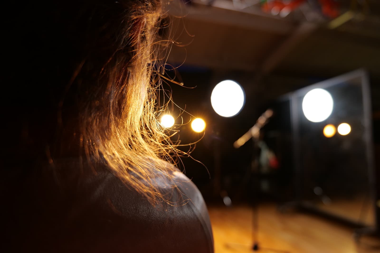 A performer seen from behind on the studio floor, hair backlit by the stage lamps in front of her.