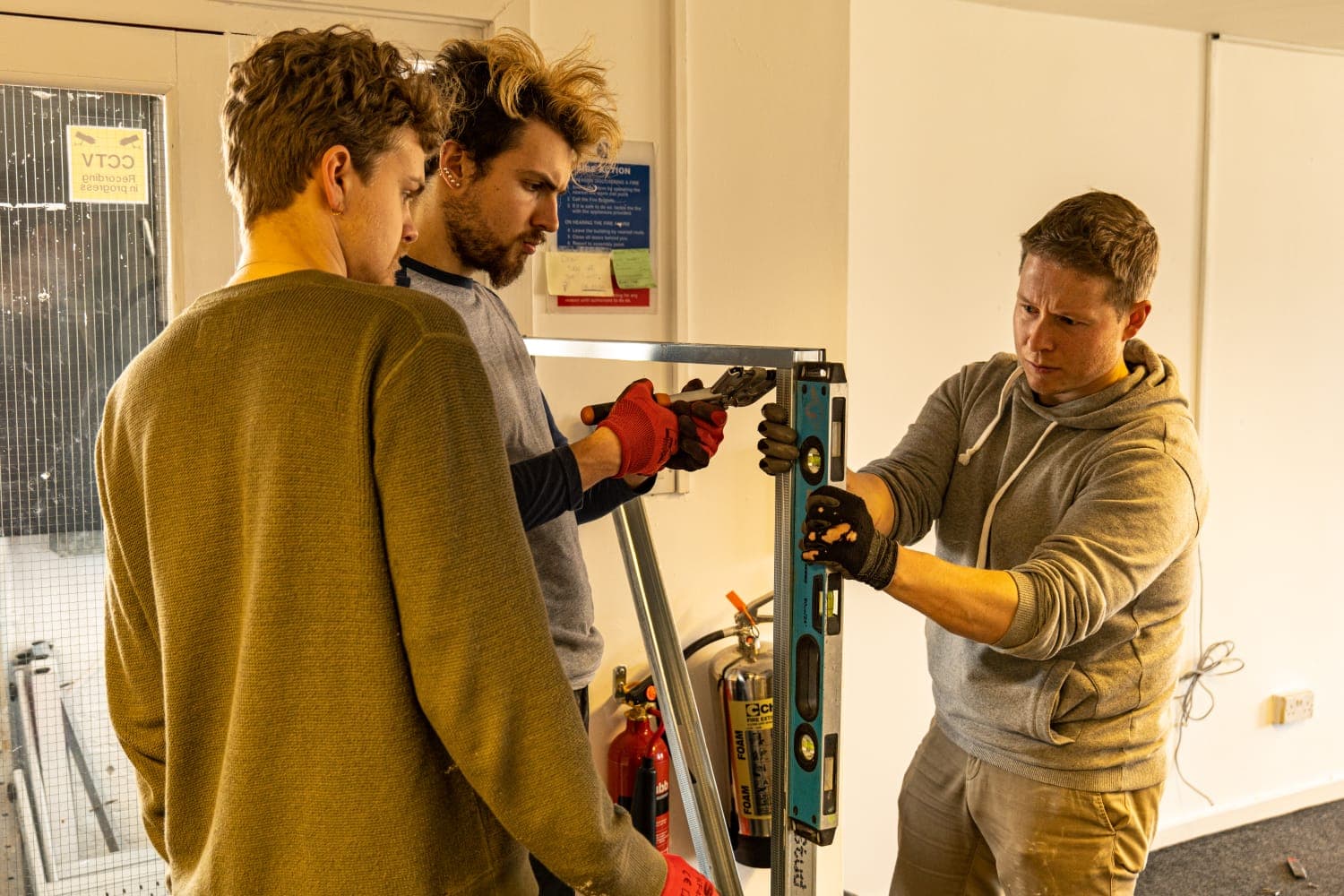Three people working with a drill to fasten some metal structure together. All concentrating and looking intently at the drill and metal work.