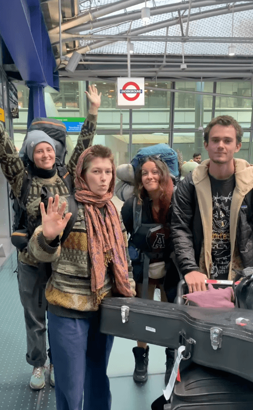 Group of four of the YIPs, travelling with backpacks and a guitar case posing and waving inside a London train station.