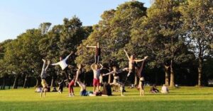 A group of people, some doing yoga, some balancing on others in the park in the sunshine