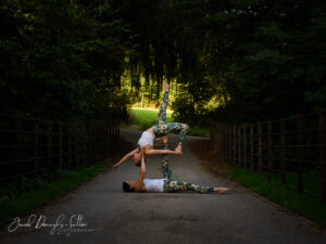 Two people performing acro yoga on a park path, with hanging willow trees overhead. One is supporting the other one in the air to strike a stretched pose, using one foot and two hands