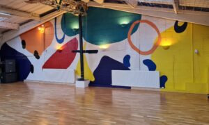A bright upstairs dance studio with polished wooden floors and a large colorful abstract mural covering one wall. The mural features bold shapes Wall-mounted lights highlight parts of the mural, giving the room a lively and creative feel.