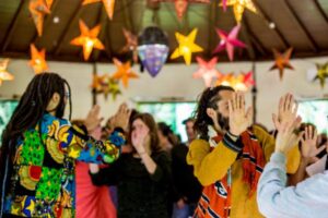 A lively group activity takes place in a decorated indoor space, with people raising their hands toward each other in pairs. Brightly colored paper star lanterns hang from the ceiling, casting a warm, festive glow. The focus is on two individuals in vibrant clothing facing each other, while the rest of the group continues in the background.