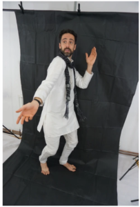 Bobak poses in a dynamic stance against a black backdrop, wearing a white kurta with matching trousers and a dark patterned scarf. His arms are extended expressively, and he looks to the side as if mid-performance.