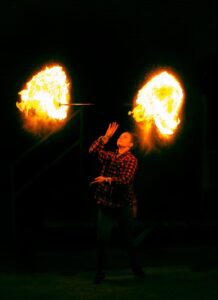 A fire performer in a red checkered shirt spins two staffs that burst into large fireballs, one on each side of them, against a dark background. The flames create bright, glowing arcs of light, illuminating the performer’s focused expression.