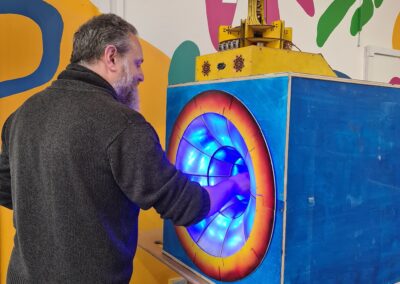 A person interacts with a brightly lit, blue-and-orange art installation at Open Source Arts. Their hand is inside a glowing tunnel of blue LED lights built into a painted wooden box, topped with a quirky yellow-and-white mechanical structure.
