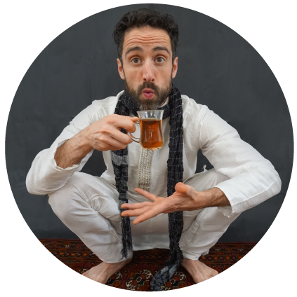 Bobak, squatting on a prayer rug, holding herbal tea while blowing on it, looking at camera.