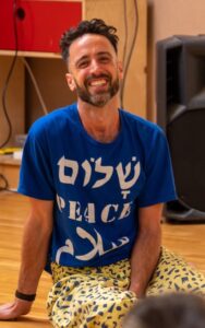 Bobak in the dance studio, sat down smiling, wearing a t-shirt saying peace, in English, Arabic and Hebrew