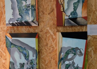 Four surreal paintings displayed together on a wooden chipboard wall at Open Source Arts. Each canvas features abstract organic shapes in muted tones of blue, grey, and green, set within geometric frames of warm reds, yellows, and oranges. One painting includes a wooden mannequin figure in an unusual pose, blending the surreal with the figurative.