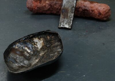 Two small sculptural objects placed on a dark grey floor. One is a pink, organic-looking cylindrical form with a rough surface, partially clasped by a curved strip of welded metal. The other is a shallow, irregularly shaped metal bowl with a scorched and textured surface, showing heat marks and welding scars.