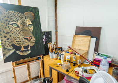 An artist’s studio with a large painting of a leopard on an easel. The desk beside it is cluttered with art supplies, including paint tubes, brushes, jars, a wooden box, a striped mug, and a bottle of water. Other canvases lean against the wall, including one with a flower and another with a figure wearing a hat. Natural light enters through a nearby window.