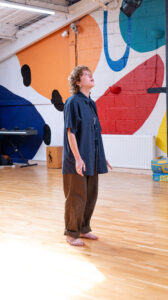 A person stands barefoot on the polished wooden floor of the Open Source Arts dance studio, looking up toward the ceiling lights. Behind them, the wall is painted with bold, colorful abstract shapes in red, blue, orange, yellow, and black.