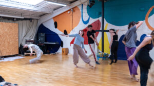 A group of dancers rehearsing in the upstairs dance studio at Open Source Arts. The wooden floor and colorful abstract mural on the wall create a bright, energetic space, with people mid-movement exploring contemporary dance.