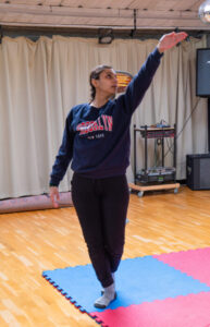 Shivani rehearsing in the build up to the performance. Practicing steps, wearing comfy clothes. She's upright. left arm high up and her head looking up towards