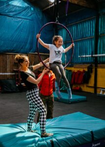 A young girl smiling as she sits in a purple aerial hoop, supported by two instructors in a training space with mats and safety equipment.