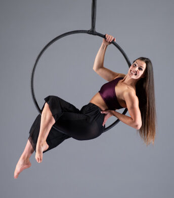 A studio photo of Jess the hoops instructors, sat and laid back in a hoop, hanging. She smiling and ready to teach hoops