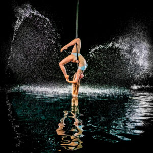 Jess performing n pole handstand, one leg hooked around the pole as water splashes dramatically behind her and reflects below.