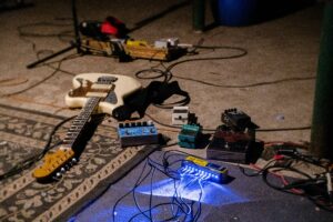 Image of an electric guitar, plus effects pedals and cables, plugged in, resting on carpet and rugs