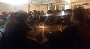 People seated around candlelit tables sharing food in a busy community dining event.