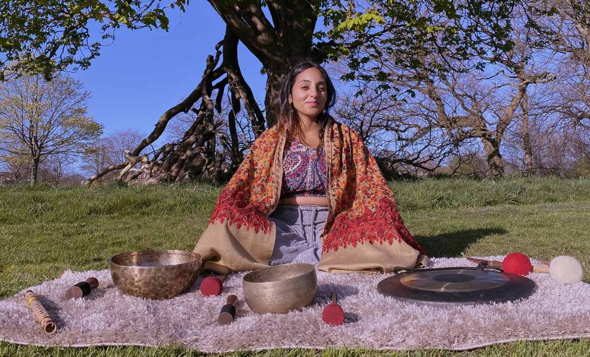Neeta seated outdoors on a blanket with singing bowls and a gong, wearing a patterned shawl.