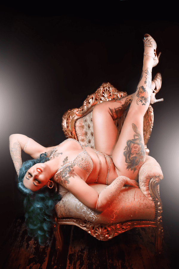 Tattooed performer with blue hair reclining upside down on an ornate chair in glamorous stagewear.